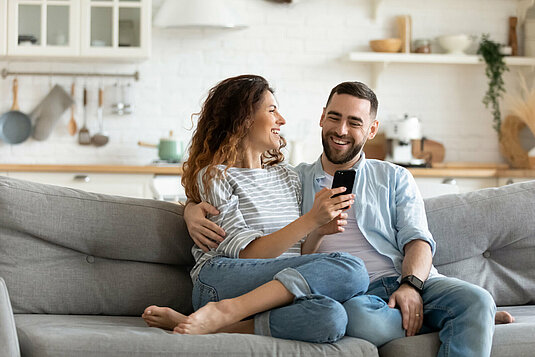 Glückliches junges Paar mit Smartphone zusammen, entspannen auf gemütliche Couch zu Hause, lächelnd überglücklich Frau und Mann umarmt, genießen Freizeit mit Gadget, sitzen auf Sofa im Wohnzimmer